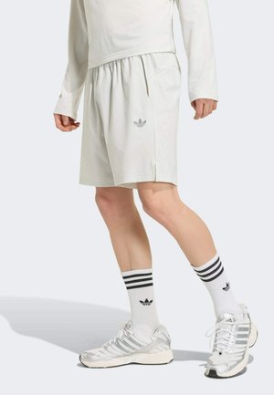 Osoba ubrana w jasnobeżowe szorty Adidas, białą bluzkę z długim rękawem, białe skarpetki Adidas i srebrne buty sportowe Adidas, stoi z jedną nogą do przodu.