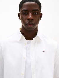 Hvit button-up skjorte laget av bomull, med en liten logo på brystet, båret over en hvit T-skjorte, med spredd krage.