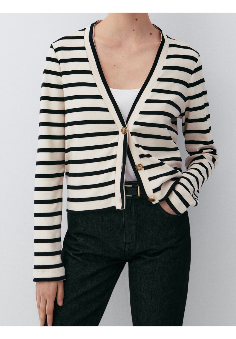 Femme portant un cardigan rayé horizontal noir et blanc avec des boutons dorés, un haut blanc et un pantalon noir, posant avec une main dans la poche.