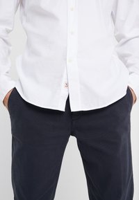 Valkoinen nappikauluspaita, joka on pistetty tummansinisten housujen sisään ja kädet taskuissa, paitahelman reunassa näkyy pieni punainen numero "3" -tägi.