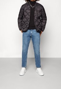 Chaqueta acolchada negra con acolchado de rombos y cuello alto sobre un suéter negro ajustado, combinada con jeans de mezclilla azul claro y zapatillas blancas.
