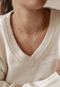 Collares en capas de oro con diseños de lunas y estrellas, adornados con una cadena delicada, llevados sobre un suéter de punto en color crema.
