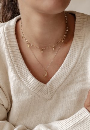 Gelaagden gouden kettingen met manen en sterren, met een delicate kettingontwerp, gedragen over een crèmekleurige gebreide trui.