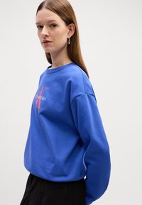 Felpa di cotone blu con collo rotondo, spalle cadute e una grafica logo rossa sul davanti. Texture liscia con una vestibilità comoda.