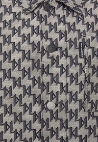 Vadderad jacka med ett grått houndstooth-mönster och mörkblå K-motiv, silverfärgade tryckknappar och en logotag på sidan.