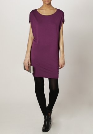 Paarse korte mouwen jurk met een losse pasvorm, gemaakt van zacht stof; gecombineerd met zwarte panty's en enkellaarsjes, een metallic clutch vasthoudend.