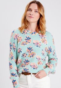 Maglione azzurro chiaro con un motivo floreale in rosa, viola e giallo. Tessuto morbido, scollo rotondo e maniche lunghe. Indossato con pantaloni bianchi.