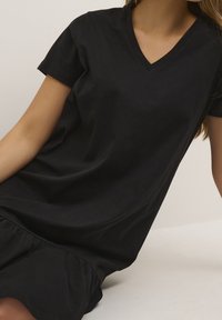 Czarna bawełniana sukienka t-shirt z dekoltem w serek, krótkimi rękawami i marszczonym dołem z falbaną. Miękka faktura i luźny krój.