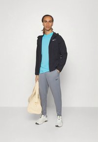 Nike Activewear-Outfit mit einer marineblauen Jacke, einem hellblauen T-Shirt, grauen Hosen und weißen Turnschuhen, dazu eine beige Sporttasche.