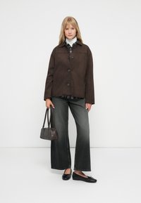 Bruine suède jas met een knoopsluiting, grijs geruit shirt eronder, donkergrijze wijde jeans en zwarte flats. Accessoire: kleine zwarte handtas.