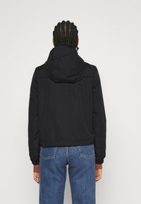 Sort hættejakke med et cropped design og elastiske manchetter. Båret over blå denim jeans, der viser en glat, mat tekstur.