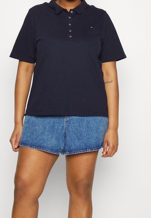 Person iført en navyblå polo-shirt med knapper og lille logo, kombineret med blå denimshorts, stående mod en ensfarvet hvid baggrund.