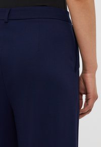 Marinblå skräddarsydda byxor med en slät textur, med bakfickor och en åtsittande midja. Utsikten visar sömmen och handens placering.