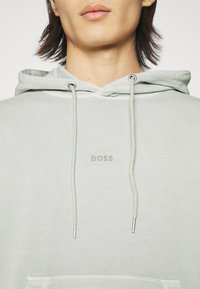 Ljusgrön hoodie med en framficka, justerbara dragskor och en subtil "BOSS"-logotyp. Tillverkad av mjukt material med en slät textur.