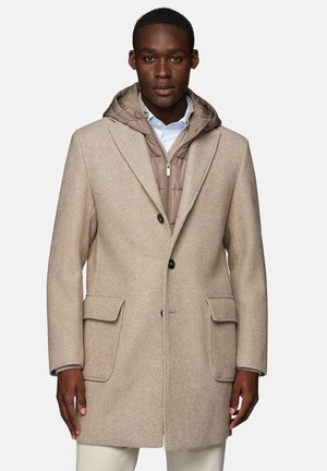 Abrigo beige de espiga con grandes bolsillos, cierre de un solo botón y forro con capucha marrón desmontable sobre una camisa blanca.