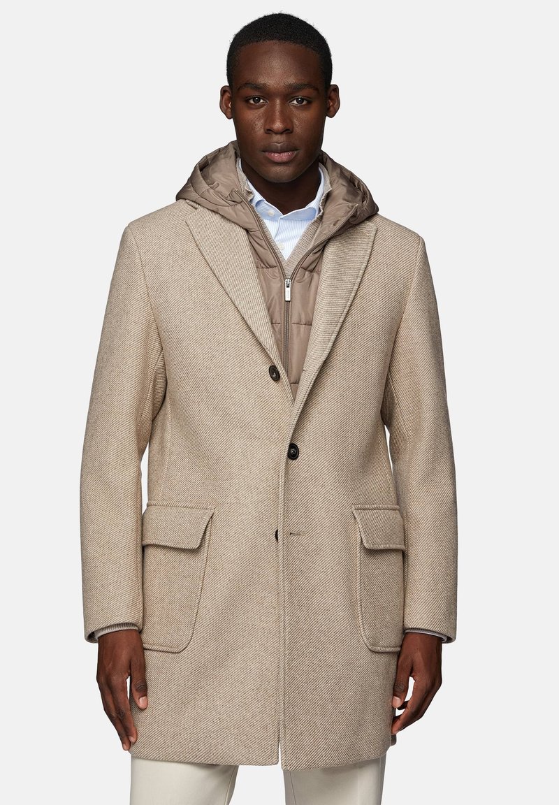 Manteau en chevrons beige avec grandes poches, fermeture à un bouton, et doublure à capuche marron amovible sur une chemise blanche.
