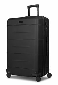 Valigia rigida nera con superficie strutturata, tre scanalature orizzontali, doppie ruote, manico retrattile e chiusura con cerniera.