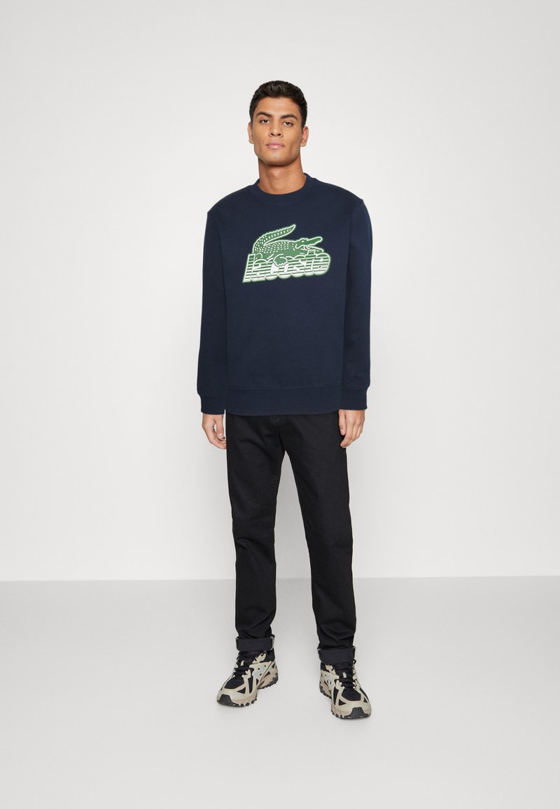 Sweatshirt bleu marine avec un logo de crocodile vert. Associé à un pantalon noir et des baskets à motifs, se tenant devant un fond uni.