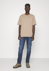 Beige cotton t-shirt, relaxed fit, round neckline; paired with blue denim jeans; wearing light gray lace-up shoes against a plain background.