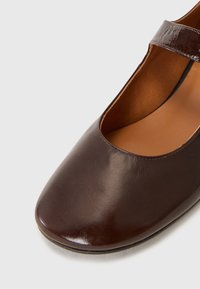 Scarpa Mary Jane in pelle marrone con punta arrotondata, tacco flat e finitura lucida. Presenta una cinghia sopra il piede per una vestibilità sicura.