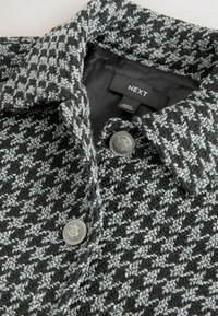 Hallide ja musta houndstooth mustriga mantel kraekaelusega, metallnuppude detailidega ja siledast voodriga. Sildil on bränd "NEXT."