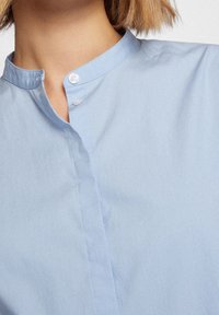 Hellblaues Button-Up-Hemd mit Stehkragen, das eine glatte Textur und einen dezenten Glanz aufweist, ergänzt durch einen einzelnen Knopfverschluss.