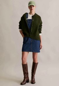 Olive green, knit cardigan over a white top with blue denim overalls, paired with a denim skirt and brown knee-high boots. Light green cap.