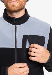Polarowa kurtka z szarym górnym panelem i czarną dolną częścią, z zamykaną na zamek kieszenią na piersi oraz wysokim kołnierzem. Miękka tekstura i casualowy design.