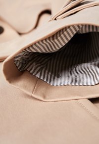 Cappotto beige con un'esterno liscio, un collo texturizzato e una fodera a righe in bianco e nero. Focalizzati sui dettagli costruttivi e sulla qualità del tessuto.