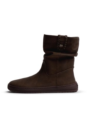 Bota de color marrón oscuro hasta la mitad de la pantorrilla con suela plana, textura de cuero suave, detalles cosidos y una pequeña anilla con un emblema metálico cerca de la parte superior.