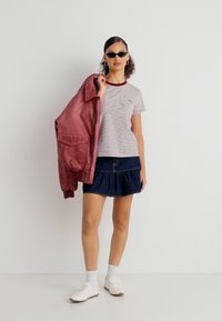 Γυναίκα που φοράει ριγέ μπλούζα, σκουρόχρωμη τζιν φούστα, λευκά αθλητικά παπούτσια και κάλτσες, κρατώντας ένα ροζ μπουφάν πάνω στον έναν ώμο, στέκεται μπροστά σε λευκό φόντο.