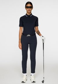 Donna in abbigliamento da golf blu marino, guanto bianco, occhiali da sole, che tiene un bastone da golf, in piedi contro uno sfondo bianco neutro.