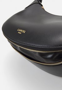 Sac en cuir noir de forme arrondie, avec une fermeture éclair dorée et un logo embossé "LANCEL PARIS". Texture lisse avec un matériel minimal.