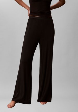 Personne portant un pantalon large noir ample et un haut noir assorti, debout pieds nus devant un fond gris uni.