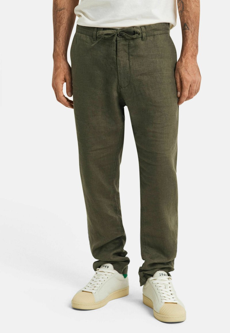 Olijfgroene linnen broek met een trekkoord en knoopsluiting, taps toelopende pasvorm, zachte textuur, in combinatie met witte sneakers met groene accenten.