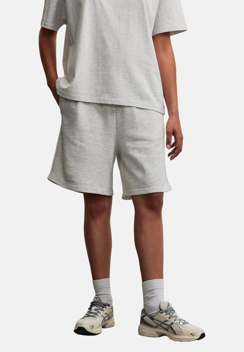 Personne portant un short gris clair, un t-shirt assorti, des chaussettes blanches hautes et des chaussures de course beige-gris, debout sur un fond uni.