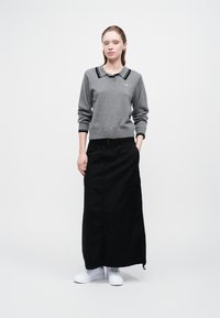 Ženska stoji oblečena v sivo dolgo-sleeve polo majico, črno krilo do gležnjev z stranskim žepom in bele superge pred enobarvno ozadju.