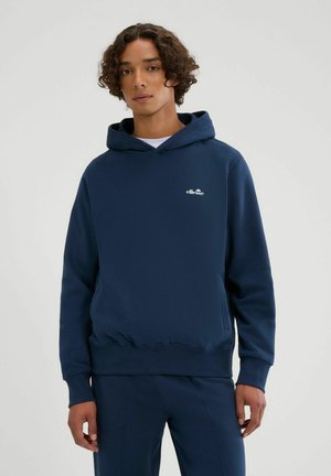 Giovane con capelli ricci indossa una felpa con cappuccio ellesse blu navy e pantaloni abbinati su uno sfondo semplice grigio chiaro.