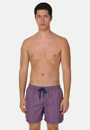 Ung mand med kort mørkt hår iført lilla badeshorts med hvidt diamantmønster og mørkeblå snøre, stående mod ensfarvet baggrund.