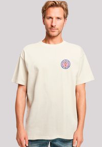 Beige cotton t-shirt with a round graphic patch on the left chest featuring a coffee cup design and text in purple and blue. Short sleeves.