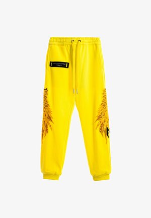 Pantalones de chándal amarillos con cintura ajustable, con herrajes negros, bolsillos laterales y diseños estilizados de alas de águila en ambas piernas.