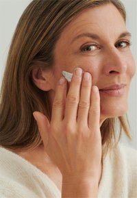 Middle-aged woman applying white cream to her cheek with her right hand, wearing a soft beige sweater, with straight brown hair.