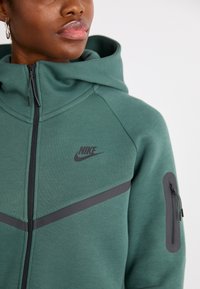 Zelená mikina Nike s kapucňou a zipsom vpredu, čiernymi akcentmi a bočnou kapsou so zipsom. Materiál vyzerá mäkko a štruktúrovane.