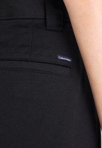 Sorte bukser laget av teksturert materiale, med en baklomme som har et lite lilla "Calvin Klein"-logo merke. Rette sømmer langs leddene.