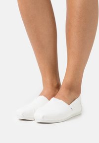 Hvite slip-on sko laget av lerret, med avrundet tå, sydd detaljer og en fleksibel såle for komfort.