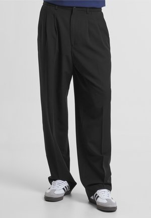 Homme portant un pantalon noir ample et des baskets blanches avec des rayures noires, debout devant un fond uni.