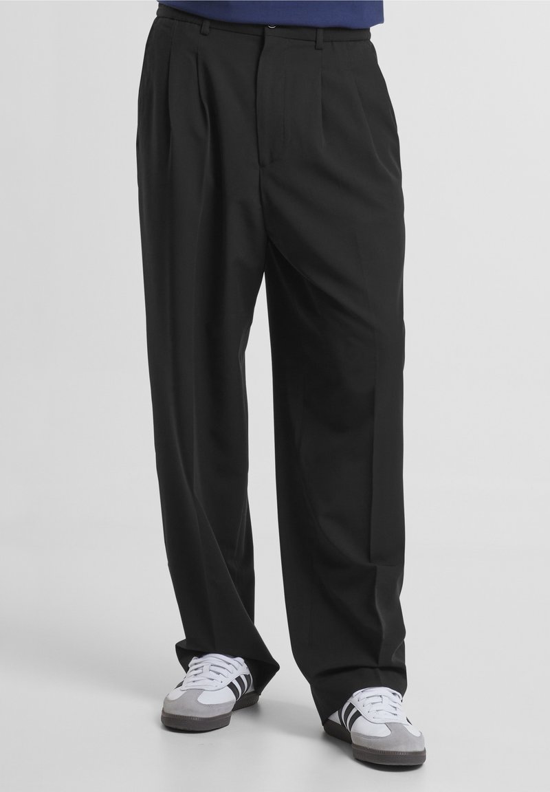Homme portant un pantalon noir ample et des baskets blanches avec des rayures noires, debout devant un fond uni.