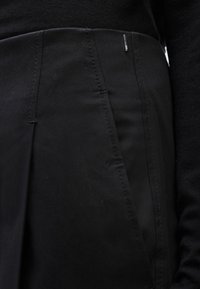 Κοντινό πλάνο ενός ατόμου που φοράει μαύρη φούστα με κάθετες ραφές και μια μικρή μεταλλική ετικέτα κοντά στη μέση, συνδυασμένη με μαύρο τοπ.