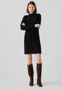 Zwarte gebreide jurk met een diamanten patroon, hoge hals en lange mouwen. Gepaard met bruin suède laarzen tot de knie.