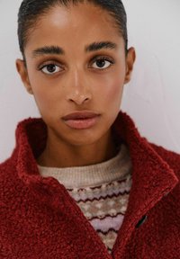 Rote Fleecejacke mit hohem Kragen, die eine strukturierte Oberfläche aufweist. Über einem cremefarbenen Pullover mit geometrischem Muster getragen.